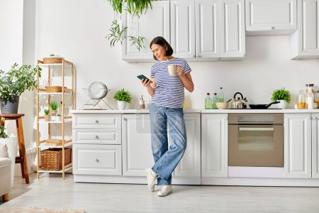 Eine reife Frau in Hauskleidung steht mit einem Handy in der Küche.