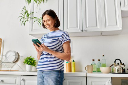 A woman in cozy homewear stands in a kitchen, holding a tablet.