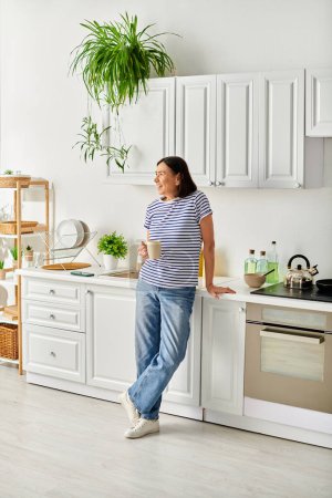 Una mujer madura en ropa de casa acogedora de pie en una cocina, junto a un fregadero.
