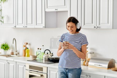 Téléchargez les photos : Femme en tenue confortable se tient dans la cuisine, absorbé dans l'écran de téléphone portable. - en image libre de droit