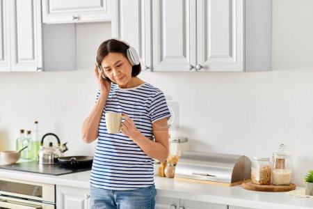 Photo for A mature woman in cozy homewear talking on a cell phone in a kitchen. - Royalty Free Image
