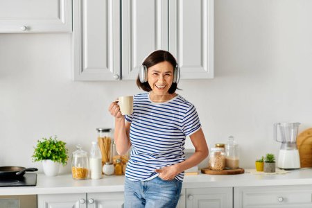 A mature woman in cozy homewear stands in the kitchen holding a cup.
