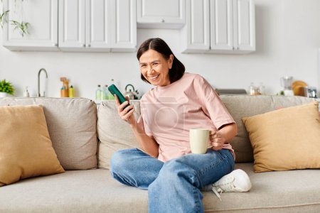 Reife Frau in kuscheliger Homewear genießt ruhigen Moment auf Couch mit Handy.