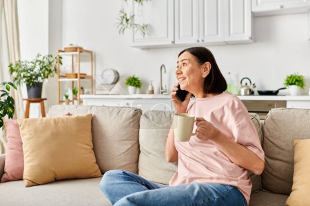 Eine reife Frau in bequemer Homewear genießt eine Tasse Kaffee, während sie auf einer Couch sitzt.