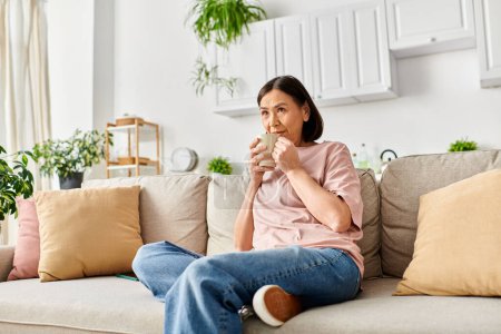 A mature woman in casual attire sits on a couch, happily eating a snack.