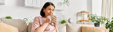 Eine reife Frau in kuscheliger Homewear sitzt auf einer Couch und nippt anmutig an einer Tasse.