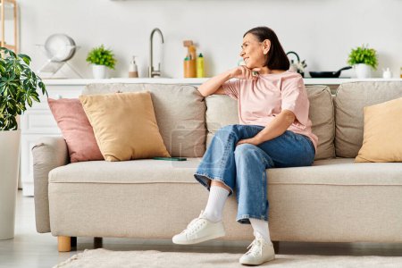 Foto de Una mujer madura y elegante vestida con ropa de casa cómoda, sentada en un sofá de felpa en una elegante sala de estar. - Imagen libre de derechos