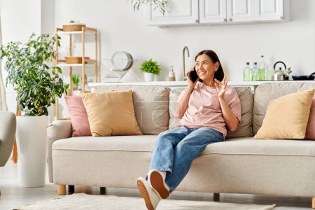 A mature woman in cozy homewear sits on a couch, engaged in a phone conversation.