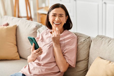 Eine reife Frau in bequemer Homewear sitzt auf einer Couch und hält ein Handy in der Hand.