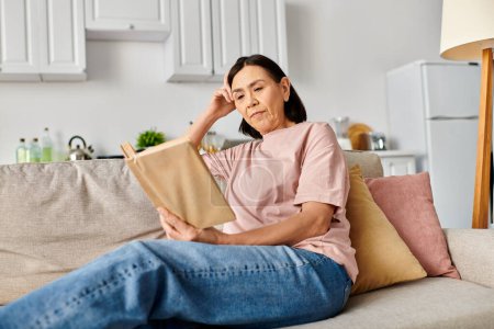 A mature woman in cozy homewear sits on a couch, engrossed in reading a book.