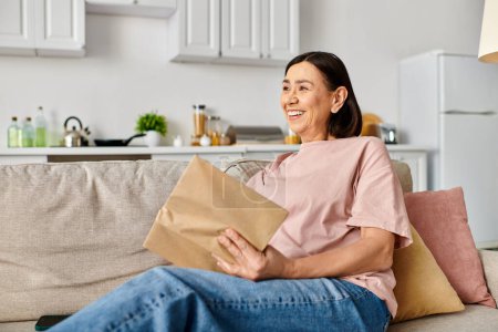 Eine reife Frau in Hauskleidung sitzt auf einem Sofa und hält eine braune Papiertüte in der Hand.