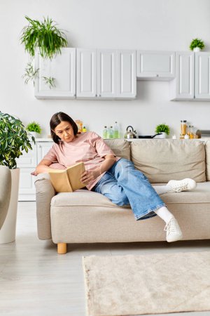 Reife Frau in bequemer Homewear, die in ein Buch vertieft ist, während sie auf einer Couch sitzt.