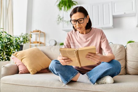 Une femme mûre en tenue confortable assise sur un canapé, absorbée par la lecture d'un livre.