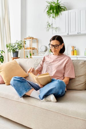 A mature woman in cozy homewear sits on a couch, fully immersed in reading a book.