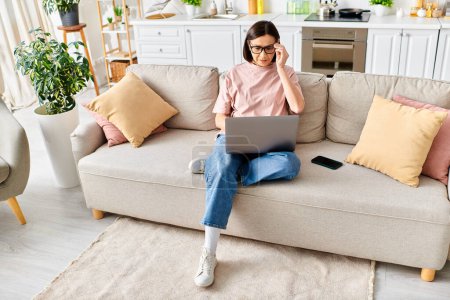 A mature woman in cozy homewear sits on a couch, focused on her laptop screen.