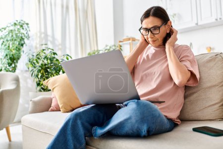 A mature woman in cozy attire using a laptop on a couch.
