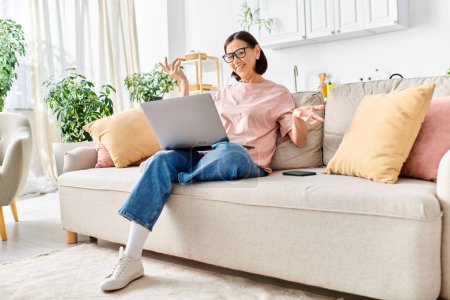 Eine reife Frau sitzt in gemütlicher Hauskleidung mit einem Laptop auf einer Couch.