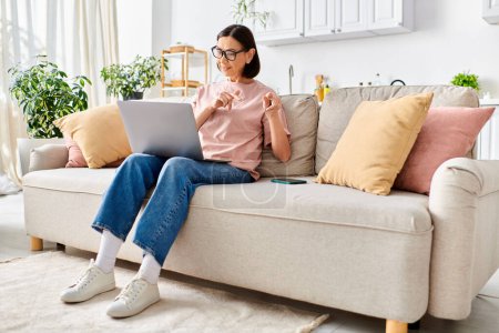 Foto de Una mujer en ropa de casa acogedora se sienta en un sofá, absorbida en su computadora portátil. - Imagen libre de derechos
