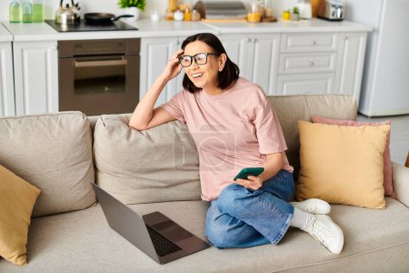 Foto de Una mujer madura en ropa de casa acogedora sentada en un sofá, trabajando en un ordenador portátil. - Imagen libre de derechos