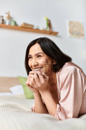 A mature woman in cozy homewear smiling on a bed.