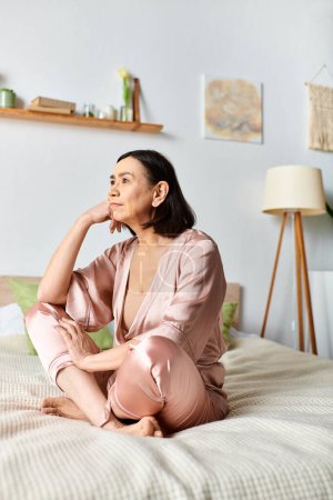 A mature woman in cozy homewear sits peacefully on a bed next to a glowing lamp.