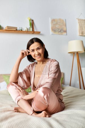 A mature woman in cozy homewear sits on a bed, smiling.