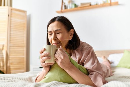 Eine Frau in gemütlicher Hausmannskleidung sitzt auf einem Bett und hält eine Tasse Kaffee in der Hand.
