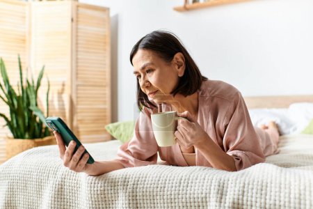 Foto de Una mujer en acogedora ropa de casa se relaja en una cama, sosteniendo una taza de café y mirando su teléfono. - Imagen libre de derechos