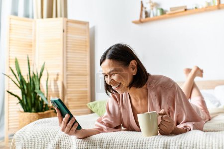 Una mujer madura en acogedores salones de ropa de casa en una cama con una taza de café y un teléfono celular.
