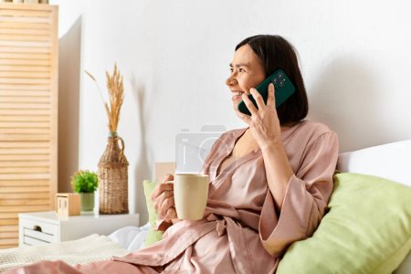 Reife Frau in kuscheliger Homewear sitzt gemütlich auf dem Bett, tief im Gespräch auf dem Handy.