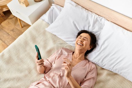 A mature woman in cozy homewear lays on a bed, holding a toothbrush.