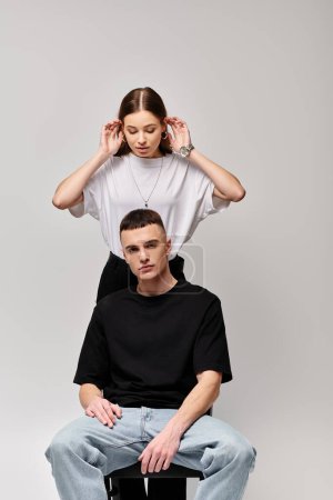 Junger Mann sitzt auf einem Frauenkopf und zeigt Gleichgewicht und Vertrauen in einer surrealen Pose vor grauem Hintergrund.
