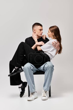 A man and a woman, a young couple in love, sitting together on a chair in a studio with a grey background.