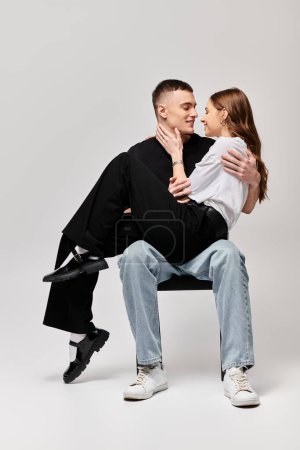Téléchargez les photos : Un jeune couple assis sur une chaise, partageant un moment intime, entouré d'un fond de studio gris. - en image libre de droit