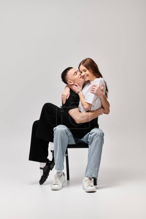 A man tenderly holds a woman in his arms while sitting on a chair in a studio with a grey background.