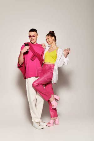 A stylish man and woman stand together, the woman in a pink outfit. The couple showcases love and fashion in a studio setting with a grey background.