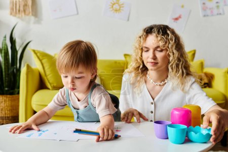 A woman with curly hair sits at a table with her toddler daughter, engaging in Montessori learning activities at home.
