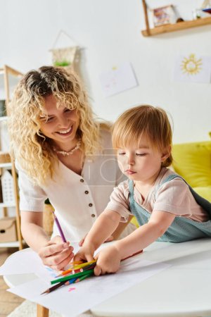 Madre de pelo rizado guía suavemente a sus hijas de niño pequeño mano mientras dibuja con lápices de colores en casa.