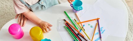 Photo for Toddler girl happily sits at a table, engrossed in play with colored pencils. - Royalty Free Image