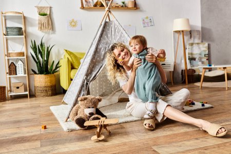 A curly-haired mother and her toddler daughter sit on the floor in front of a teepee tent, engaging in the Montessori method of education at home.