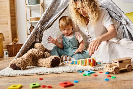 Foto de Curly mother and toddler daughter engaging in imaginative play inside a colorful play tent in a cozy home setting. - Imagen libre de derechos