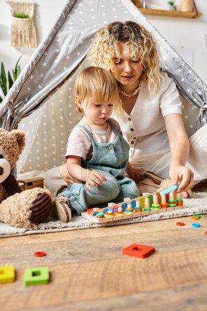 Eine lockige Mutter und ihre kleine Tochter spielen fröhlich in der Nähe eines Teddybären und üben sich in fantasievoller Montessori-inspirierter Erziehung zu Hause.