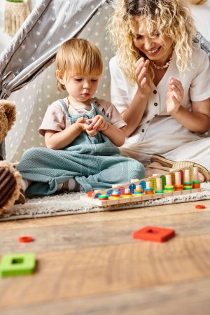 Eine lockige Mutter und ihre spielfreudige Kleinkindtochter beim Binden eines Teddybären nach Montessori-Methode zu Hause.