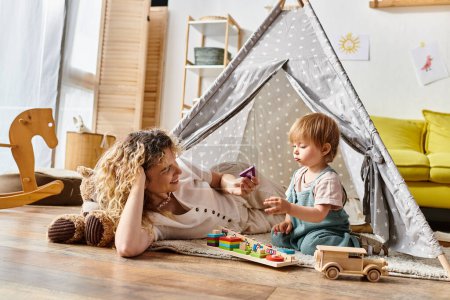 Lockige Mutter und Kleinkind-Tochter spielen fröhlich zusammen in einem bunten Spielzelt und umarmen die Montessori-Pädagogik zu Hause.