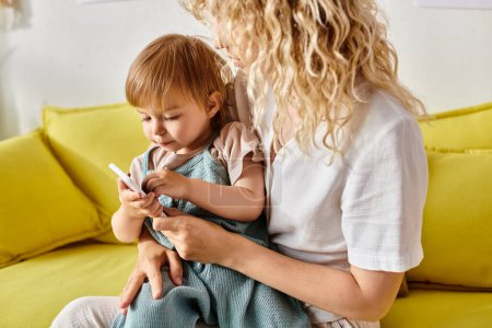 Curly-haired mother holding toddler daughter who is mesmerized by a cell phone screen at home.