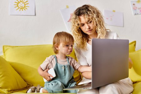 A curly-haired mother and her toddler daughter sit on a couch, engrossed in a laptop screen.
