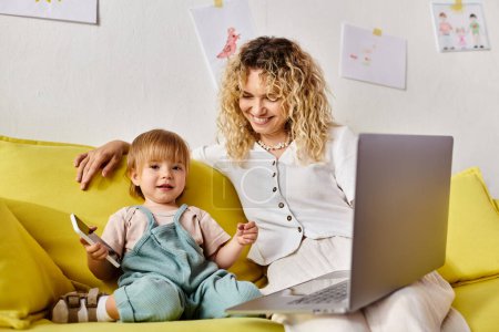 Foto de Una madre de pelo rizado se sienta en un sofá amarillo con su hija pequeña, ambos absortos en un ordenador portátil. - Imagen libre de derechos