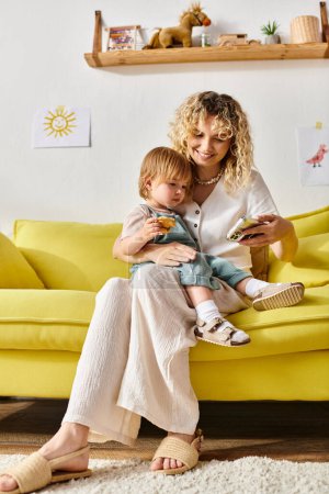 Foto de Una madre de pelo rizado se sienta en un sofá amarillo, sosteniendo a su hija en un cálido y cariñoso abrazo en casa. - Imagen libre de derechos