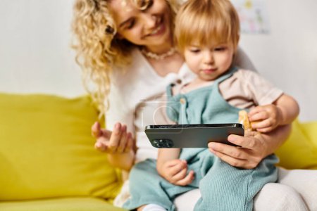 A curly mother attentively holds her toddler daughter while looking at a tablet in a cozy home setting.