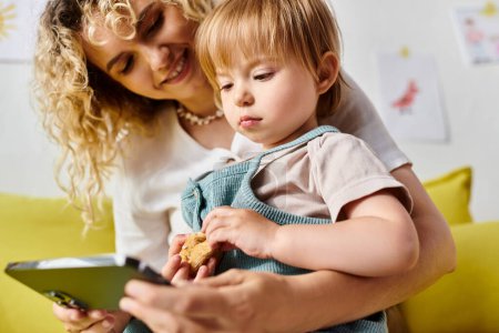 Eine lockige Mutter hält ihre kleine Tochter in der Hand, während sie in einer gemütlichen häuslichen Umgebung auf das Smartphone schaut.